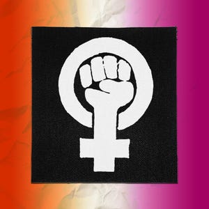 May include: A square black fabric patch featuring a white screen-printed graphic of a raised fist centered within a female gender symbol. The design is bold and high-contrast, with the white ink clearly visible against the dark black textile background.