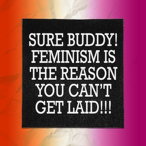May include: A square black fabric patch featuring white text that reads: SURE BUDDY! FEMINISM IS THE REASON YOU CAN'T GET LAID!!! The lettering is presented in a bold, sans-serif font, centred on the patch with a visible woven fabric texture.