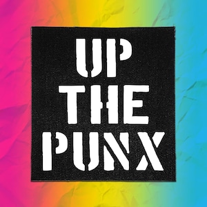 May include: A square black fabric patch featuring the white text UP THE PUNX in a bold, stencil-style font. The lettering is centered on the patch, which has a visible woven texture throughout the black background and the white printed letters.