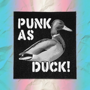 May include: A square black fabric patch featuring a white halftone graphic of a duck in profile. The text PUNK AS DUCK! is printed in a bold, white, distressed stencil-style font, with the words positioned above and below the duck illustration.