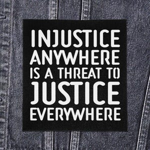May include: A black square patch with white text that reads: "INJUSTICE ANYWHERE IS A THREAT TO JUSTICE EVERYWHERE." The patch is sewn onto a denim jacket.