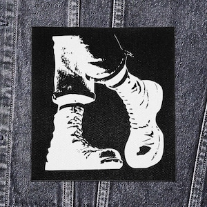 Op de afbeelding: Zwart-wit patch met een gestileerde illustratie van benen en voeten in combat boots en gestreepte sokken. De vierkante patch is geplaatst op een spijkerjasje. Grafisch ontwerp.
