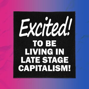 May include: A square black fabric patch featuring white text that reads Excited! TO BE LIVING IN LATE STAGE CAPITALISM! The patch has a visible woven texture and clean, straight edges, presenting a bold, high-contrast design for textile application.