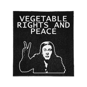 Può includere: Una toppa quadrata in tessuto nero con un'illustrazione grafica bianca di una persona con i capelli lunghi che fa il segno di pace. Sopra la figura, il testo VEGETABLE RIGHTS AND PEACE è stampato in un carattere bianco, grassetto e senza grazie.