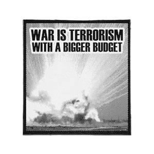 Pode incluir: Um emblema quadrado em preto e branco com um gráfico de meio-tom de uma explosão. O texto WAR IS TERRORISM WITH A BIGGER BUDGET está impresso em uma fonte sem serifa em negrito dentro de uma caixa retangular branca no topo do emblema.