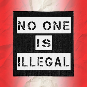 May include: A square fabric patch with a black background and two horizontal white rectangular bands. The text NO ONE is printed in black stencil-style font on the top band, IS is centred in the middle, and ILLEGAL is on the bottom band.