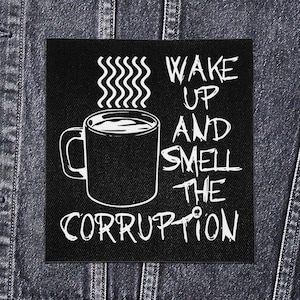 May include: Black square patch featuring a white coffee mug graphic with steam and the words "WAKE UP AND SMELL THE CORRUPTION". The patch is likely fabric and is shown on a denim jacket.