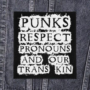 May include: A black square patch with white text that reads "PUNKS RESPECT PRONOUNS AND OUR TRANS KIN." The patch is on a denim jacket. The text has a distressed, grunge-style appearance.
