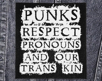 May include: A black square patch with white text that reads "PUNKS RESPECT PRONOUNS AND OUR TRANS KIN." The patch is on a denim jacket. The text has a distressed, grunge-style appearance.