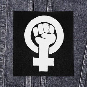 以下が含まれることがあります： 黒い正方形のパッチ。白いグラフィックは、円の中の握りこぶしと女性のシンボルです。パッチはおそらく布製で、デニムの背景に配置されています。