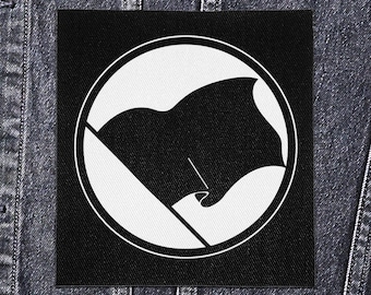May include: Black and white patch featuring a black flag design within a white circle. The patch is square and set against a dark denim jacket. The flag has a curved shape, and the design is simple and bold.