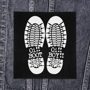 Peut inclure: Écusson carré noir avec deux empreintes de bottes blanches et les mots "Oi!! BOOT BOY!!" en blanc. L'écusson est présenté sur un fond de veste en jean.