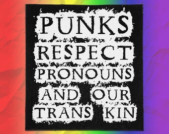 Op de afbeelding: Een vierkante zwarte stoffen patch met witte tekst in een verweerde stijl die luidt: PUNKS RESPECT PRONOUNS AND OUR TRANS KIN. De letters zijn gerangschikt in vijf regels met een ruw, getextureerd uiterlijk tegen de zwarte achtergrond.