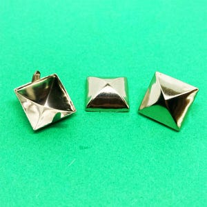 Può includere: Tre borchie piramidali in metallo color oro con una finitura liscia e riflettente. Le borchie sono di forma quadrata con cime appuntite. Una borchia è posizionata per mostrare il lato inferiore cavo e i rebbi, mentre le altre due mostrano la vista dall'alto.