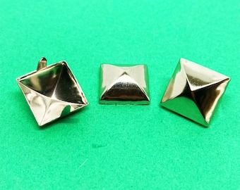 May include: Three metallic gold-colored pyramid studs with a smooth, reflective finish. The studs are square-shaped with pointed tops. One stud is positioned to show its hollow underside and metal prongs, while the other two show the top view.