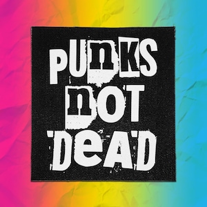 Op de afbeelding: Een vierkante zwarte stoffen patch met de witte tekst PUNKS NOT DEAD in een vet, verweerd bloklettertype. De letters hebben een ruw, getextureerd uiterlijk met zichtbare witte spikkels en grillige randen tegen de zwarte achtergrond.