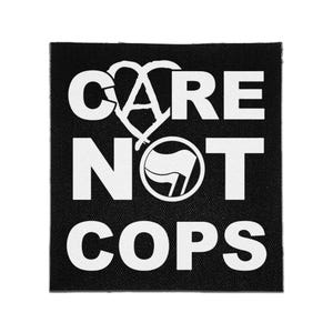 Op de afbeelding: Een vierkante zwarte stoffen patch met de witte tekst CARE NOT COPS. De letter A in CARE is vervangen door een anarchistisch symbool en de letter O in NOT is vervangen door een cirkelvormig icoon met een vlagsymbool in wit en zwart.