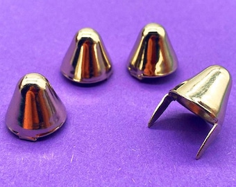 May include: Four metallic gold-toned cone-shaped studs with a smooth, reflective surface. Each stud features a rounded top and a wide base. One stud is positioned on its side, revealing two thin, pointed metal prongs extending from the bottom.