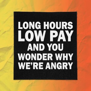 May include: A square black fabric patch featuring white text that reads: LONG HOURS LOW PAY AND YOU WONDER WHY WE'RE ANGRY. The patch has a visible woven texture and is set against a background that transitions from yellow to orange.