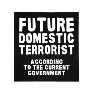 May include: A square black fabric patch featuring bold white text that reads FUTURE DOMESTIC TERRORIST ACCORDING TO THE CURRENT GOVERNMENT. The design is printed in a clean, sans-serif font on a textured material with visible woven fabric details.