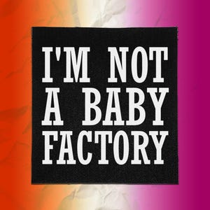 Puede incluir: Un parche de tela cuadrado de color negro con texto bordado en blanco que dice I'M NOT A BABY FACTORY en una fuente negrita sin remates. El parche tiene una textura tejida visible y bordes rectos y limpios, diseñado para ropa o accesorios.