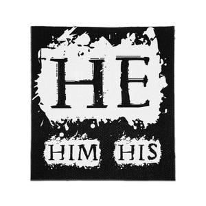 May include: A square black fabric patch featuring a white distressed ink design. The large letters HE are centered at the top, with the words HIM and HIS positioned below in smaller text, all set against a textured, splattered white background.