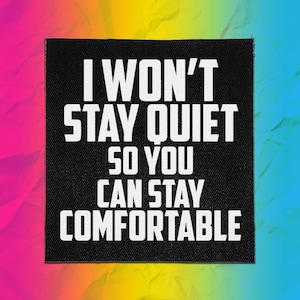 Puede incluir: Un parche cuadrado de tela negra con texto blanco que dice I WON'T STAY QUIET SO YOU CAN STAY COMFORTABLE. El parche tiene una textura tejida visible y presenta letras mayúsculas negritas sin remates centradas sobre el fondo negro.