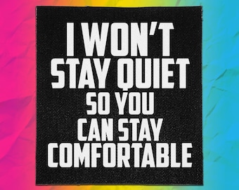 Op de afbeelding: Een vierkante zwarte stoffen patch met witte tekst die luidt I WON'T STAY QUIET SO YOU CAN STAY COMFORTABLE. De patch heeft een zichtbare geweven textuur en is voorzien van dikke, schreefloze hoofdletters gecentreerd op de zwarte achtergrond.