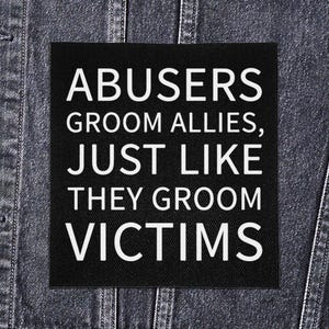 Op de afbeelding: Een zwart vierkant patch met witte tekst die luidt: "ABUSERS GROOM ALLIES, JUST LIKE THEY GROOM VICTIMS." De patch is geplaatst tegen een denim jas achtergrond.