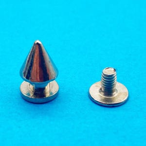 Könnte beinhalten: Ein kegelförmiger Nietverschluss aus silberfarbenem Metall, bestehend aus zwei separaten Teilen. Ein Teil hat eine spitze, kegelförmige Oberseite auf einer flachen, runden Basis, das zweite Teil ist eine flache, runde Basis mit Gewindeschraube.