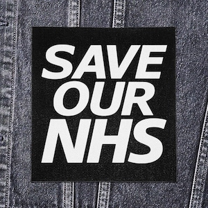 May include: A black square patch with the white text "SAVE OUR NHS". The patch is displayed on a denim jacket. The text is in a bold, sans-serif font.