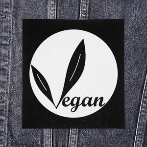 Op de afbeelding: Zwart vierkantige patch met een witte cirkel en twee gestileerde bladvormen. Het woord "Vegan" staat in een cursief lettertype eronder. De patch wordt getoond op een spijkerjasje.