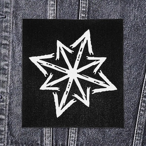 Può includere: Una toppa quadrata nera con un disegno a stella bianca composto da frecce rivolte verso l'esterno. La toppa è probabilmente in tessuto e viene mostrata su una giacca di jeans.