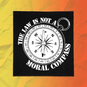 May include: A square black fabric patch featuring a white graphic of a circular compass with directional markers and degree increments. The text THE LAW IS NOT A MORAL COMPASS is printed in white capital letters around the compass illustration.