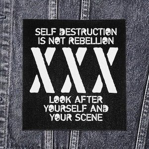 以下が含まれることがあります： 白い文字で「SELF DESTRUCTION IS NOT REBELLION」と書かれた黒い正方形のパッチ。大きな白い「XXX」の文字の上にあり、「LOOK AFTER YOURSELF AND YOUR SCENE」が下にあります。パッチはデニムジャケットにあります。