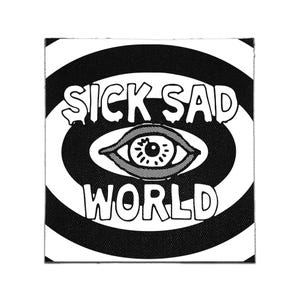 Op de afbeelding: Een vierkante stoffen patch met een zwart-wit grafisch ontwerp. Het midden toont een gestileerd oog omringd door concentrische ovale ringen. De tekst SICK SAD WORLD is gedrukt in een vet, handgetekend lettertype boven en onder het centrale oog.