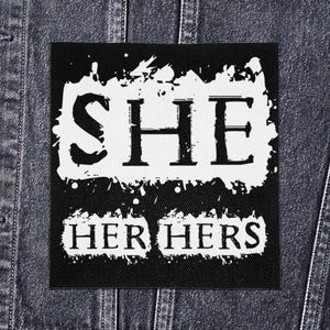 Op de afbeelding: Zwart-witte patch met de woorden "SHE" boven "HER HERS". De tekst is vetgedrukt, met een distressed lettertype, met een witte achtergrond en zwarte rand. De patch zit op een spijkerjasje.