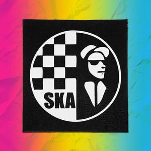 May include: A square black fabric patch featuring a white circular design. The design includes a checkered pattern on the left, the word SKA in bold capital letters at the bottom, and a stylized silhouette of a person wearing a hat and sunglasses.