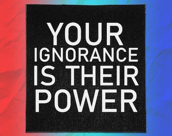 May include: A square black fabric patch featuring the white text YOUR IGNORANCE IS THEIR POWER in a bold, sans-serif font. The patch has a visible woven texture and clean, straight edges, presented against a vibrant red and blue gradient background.