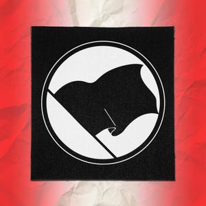 May include: A square black fabric patch featuring a white circular border enclosing a stylised black flag graphic. The design includes a diagonal white line extending from the bottom left to the top right, intersecting the circular frame.