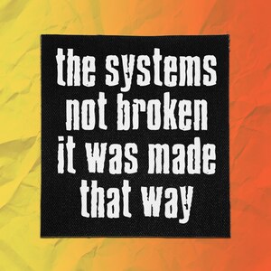 Könnte beinhalten: Ein quadratischer schwarzer Stoffaufnäher mit weißem Text, der lautet: the systems not broken it was made that way. Die Schrift ist in einer fetten, serifenlosen Schriftart gehalten und wirkt vor dem dunklen Hintergrund leicht texturiert.