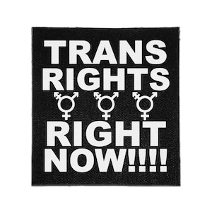 Puede incluir: Un parche de tela negro cuadrado con el texto blanco TRANS RIGHTS RIGHT NOW!!!! en letras mayúsculas negritas. Entre las dos líneas de texto hay tres símbolos blancos que representan la identidad de género trans, cada uno con una cruz y una flecha.