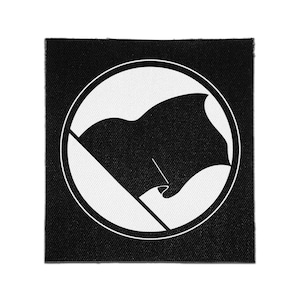 May include: A square fabric patch featuring a black and white design. The graphic consists of a solid black flag waving on a pole, enclosed within a white circular border, all set against a solid black square background with a visible fabric texture.