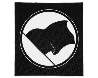 May include: A square fabric patch featuring a black and white design. The graphic consists of a solid black flag waving on a pole, enclosed within a white circular border, all set against a solid black square background with a visible fabric texture.
