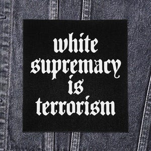 Peut inclure: Un écusson carré noir avec des lettres gothiques blanches qui se lisent "white supremacy is terrorism". L'écusson est affiché sur une veste en jean.