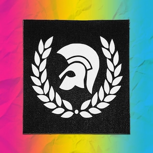 May include: A square black fabric patch featuring a white graphic of a Spartan helmet centered within a circular laurel wreath. The design is printed in a clean, bold style on a textured woven material, creating a high-contrast visual element.