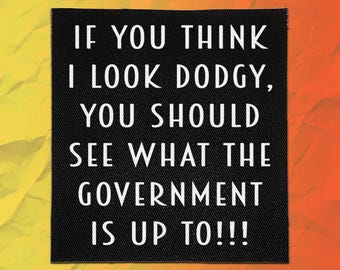 Op de afbeelding: Een vierkante zwarte stoffen patch met witte tekst: IF YOU THINK I LOOK DODGY, YOU SHOULD SEE WHAT THE GOVERNMENT IS UP TO!!! De patch heeft een zichtbare geweven textuur en is geplaatst tegen een heldere gele en oranje achtergrond.
