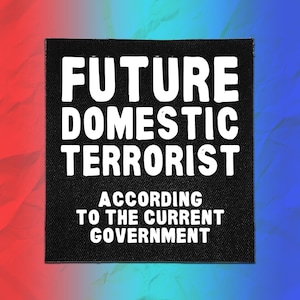 May include: A square black fabric patch featuring white text that reads FUTURE DOMESTIC TERRORIST ACCORDING TO THE CURRENT GOVERNMENT. The lettering is bold and sans-serif, centered on the patch, which has a visible woven texture throughout.