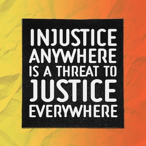 May include: A square black fabric patch featuring the white text INJUSTICE ANYWHERE IS A THREAT TO JUSTICE EVERYWHERE. The lettering is in a bold, sans-serif font, and the patch has a visible woven texture throughout the black background surface.