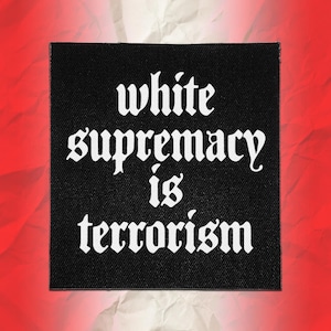 Könnte beinhalten: Ein quadratischer schwarzer Stoffaufnäher mit weißem Text in einer gotischen Schriftart, der "white supremacy is terrorism" lautet. Der Aufnäher hat eine sichtbare Webstruktur und ist vor einem leuchtend roten Hintergrund mit Knittereffekt zentriert.